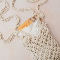 A cream macrame shoulder bag with a white fabric insert and a small orange paper tag, laid flat on a light pink background.
