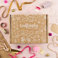 A Craftosity Craft Kits subscription box in brown kraft paper with white line drawings of crafting tools, surrounded by craft supplies like ribbon, scissors, thread, and pom-poms on a pink background.
