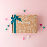 A flat-lay product shot of a Crafttivity Craft Kits box on a solid pink background, decorated with a teal velvet ribbon and bow. The box is wrapped in kraft paper with white line drawings of various craft tools, and is surrounded by small colorful Christmas ornaments.
