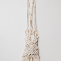 An off-white macrame shoulder bag with a long woven strap and fringed bottom, hanging against a plain light gray background.