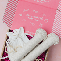An open pink gift box filled with baby craft supplies, including two rolled cream cord tubes, a small cream fabric pouch, wooden rings, and a pink shred lining. The inside lid reads 'Have a wonderful Craftmoon!' and shows sewing-themed illustrations.