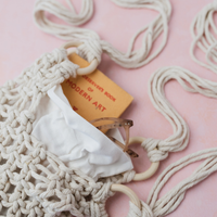 A cream macrame shoulder bag with a chunky woven texture and wooden ring handles, shown on a soft pink background with travel accessories peeking out from inside.